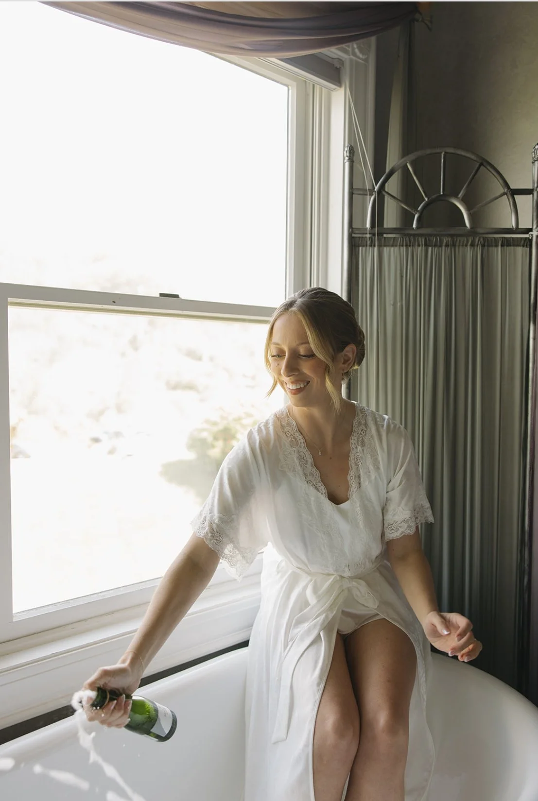 A woman in a white lace robe sitting on a bathtub, holding a bottle of champagne, smiling joyfully, with a window and curtain in the background.