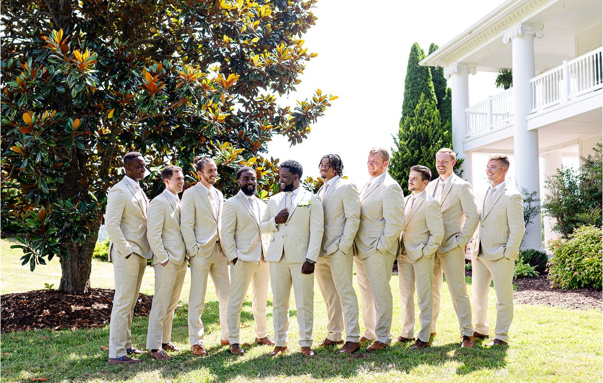A group of ten men in cream-colored suits standing outdoors in front of a large tree and a white building, engaged in conversation and smiling.