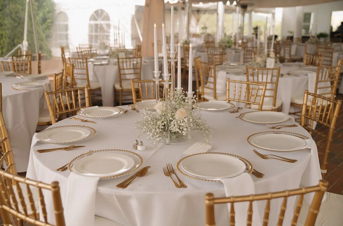 A banquet hall set up for a formal event with round tables, gold Chiavari chairs, white tablecloths, white plates with gold beaded rims, gold flatware, and a centerpiece of white flowers and tall white candles.