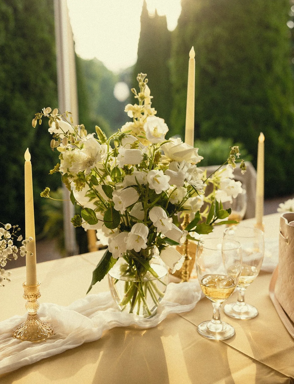A table with a floral centerpiece of white flowers in a glass vase, surrounded by three candles, three glasses of wine, and a beige cloth runner, with greenery visible outside the window in the background.