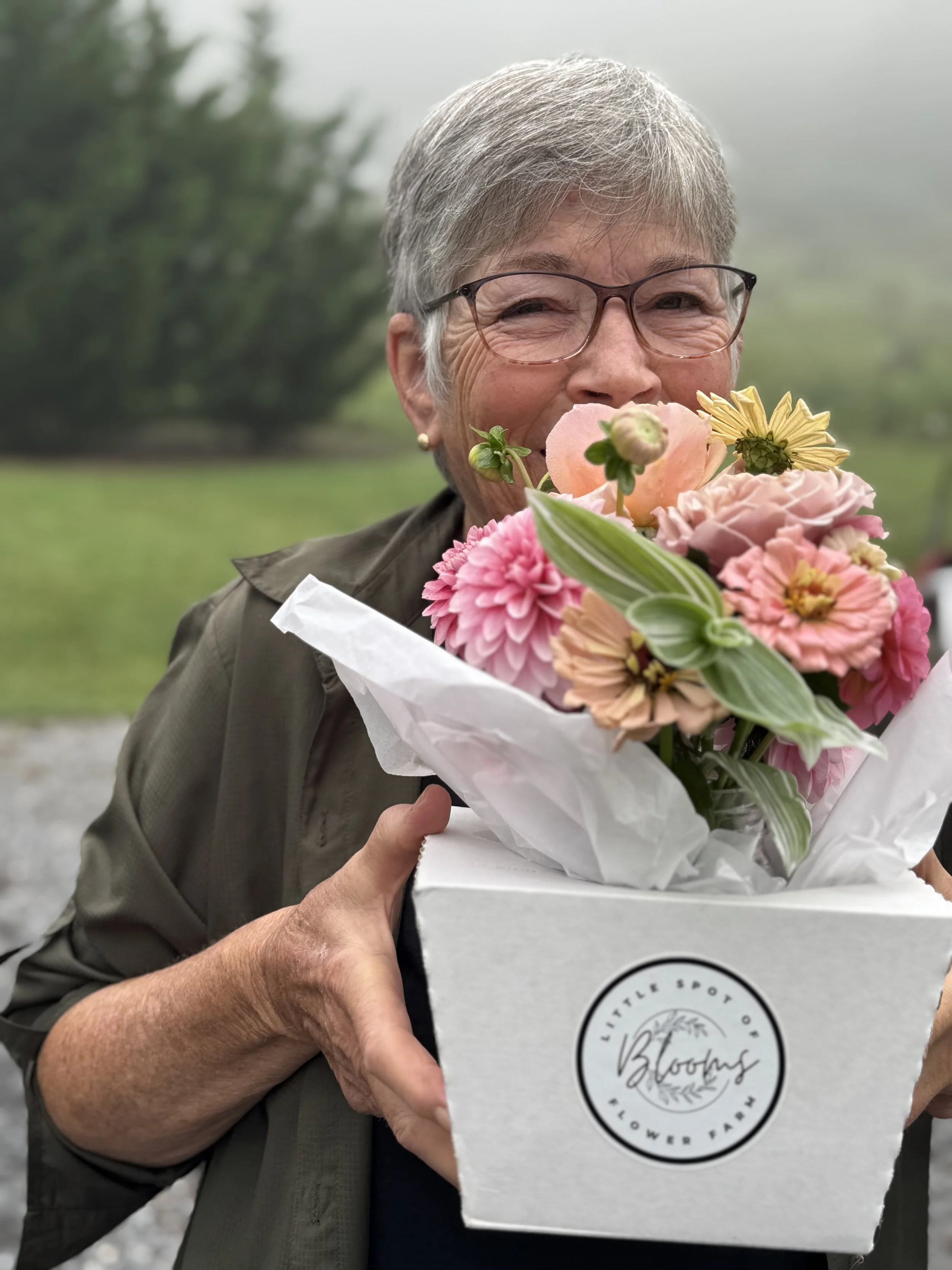 An elderly woman with short gray hair and glasses smiling while holding a bouquet of pink, peach, and yellow flowers in a white box labeled "Little Spot of Blooms Flower Farm." She is outdoors with a blurred green landscape in the background.