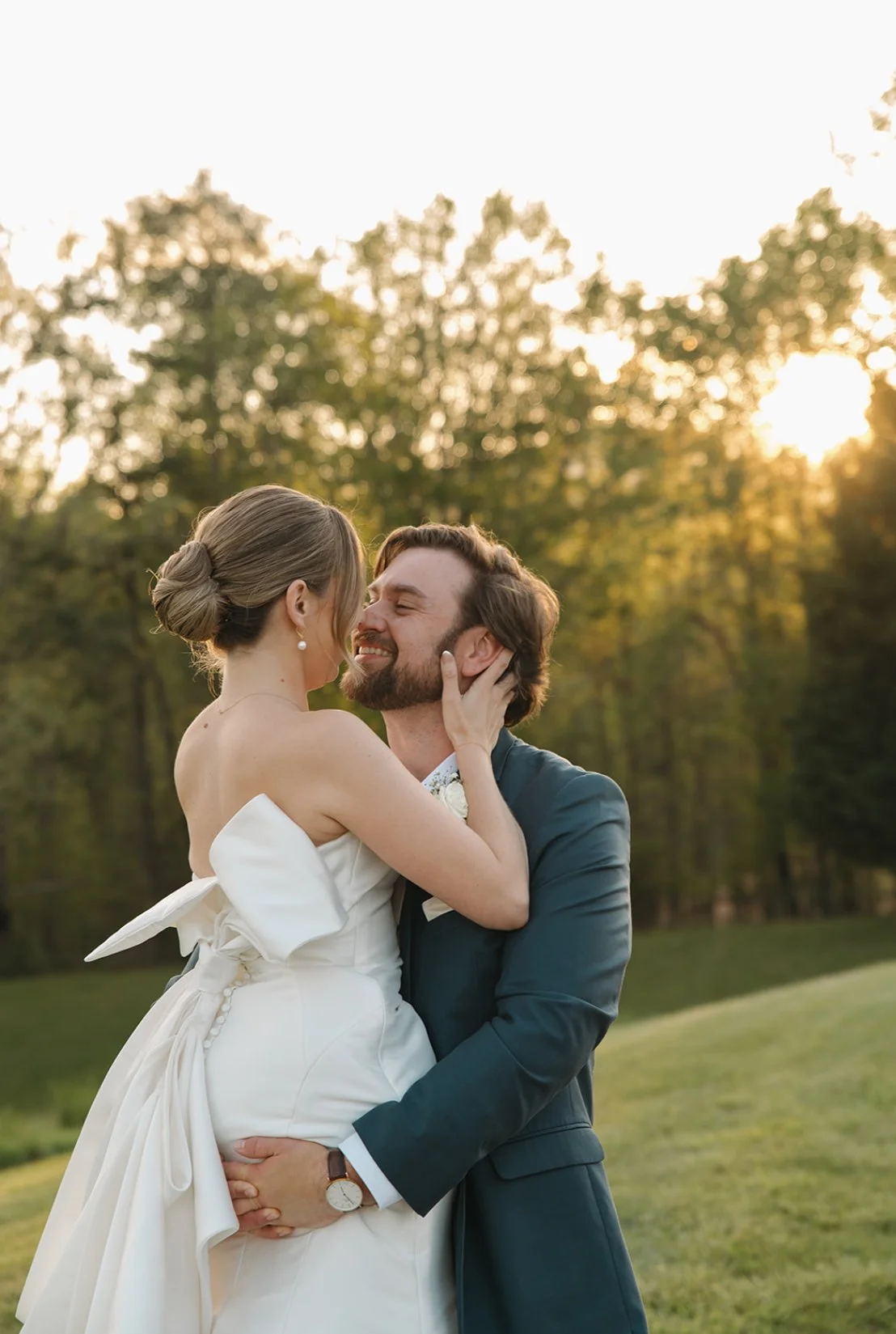 A bride and groom embrace outdoors at sunset, smiling and holding each other with trees in the background.