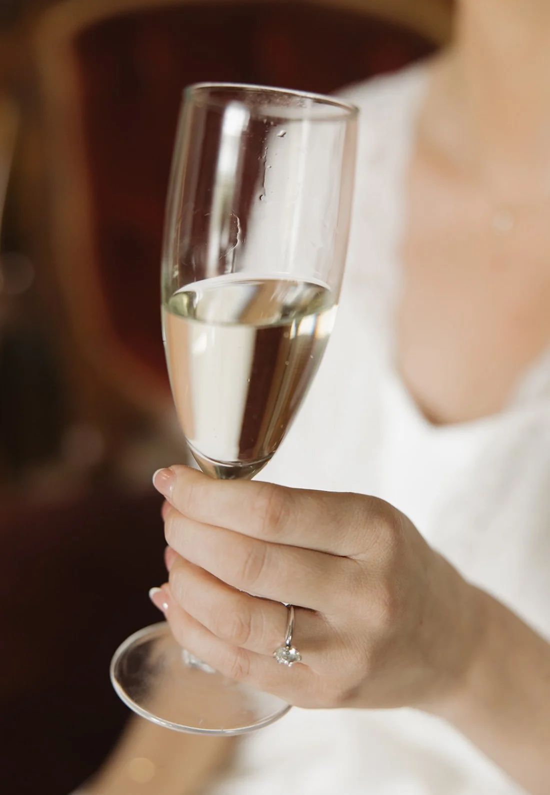 A person holding a champagne flute filled with champagne, wearing a ring on their finger, with a blurred background.