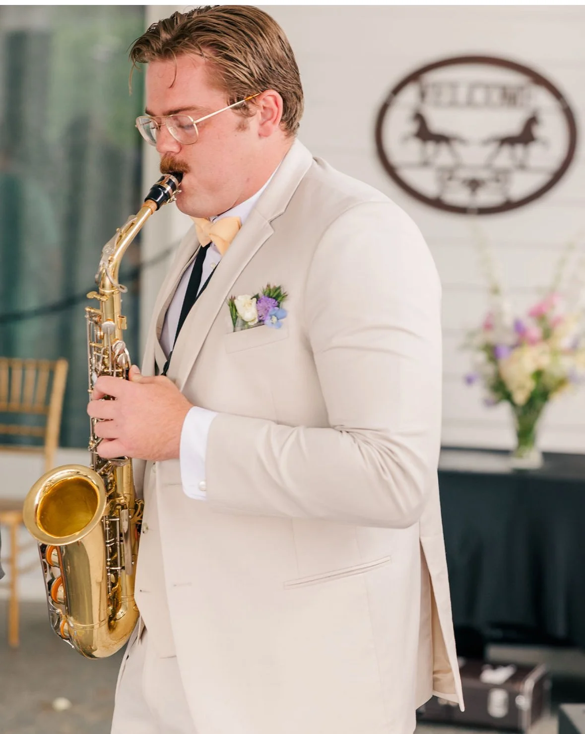 A man with glasses and a mustache, dressed in a white tuxedo with a yellow bow tie, playing a saxophone at an event. There is a blurred sign and a flower arrangement in the background.