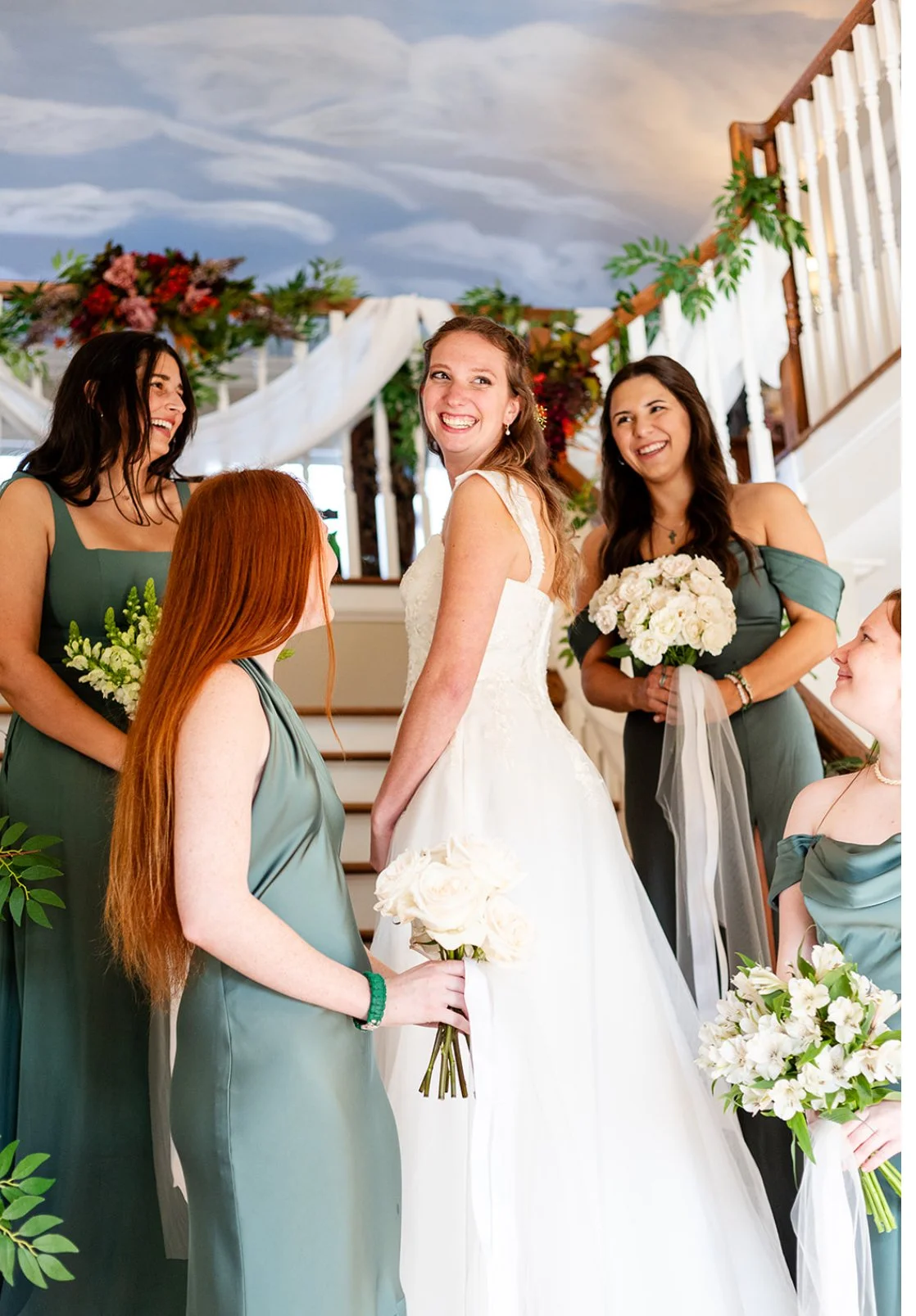 A bride and her bridesmaids smiling and holding bouquets during a wedding celebration inside a decorated venue.