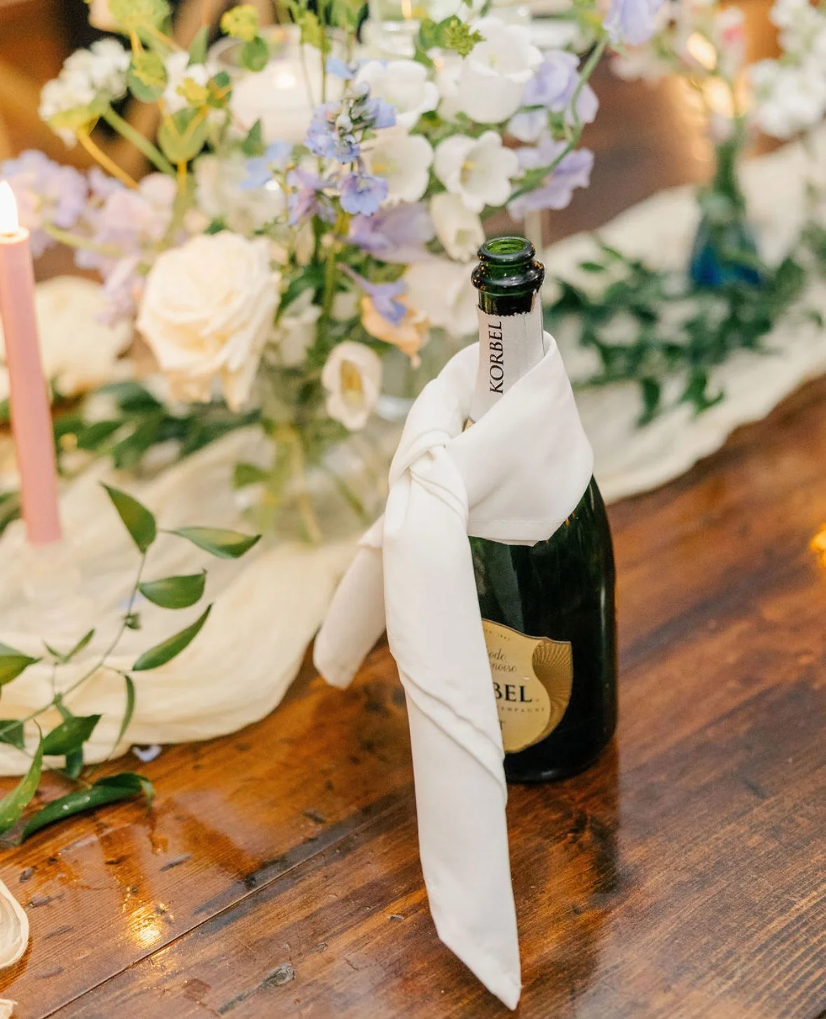 A wine bottle on a wooden table with a white cloth tied around its neck, part of a floral centerpiece with white and light purple flowers, and pink candles in the background.