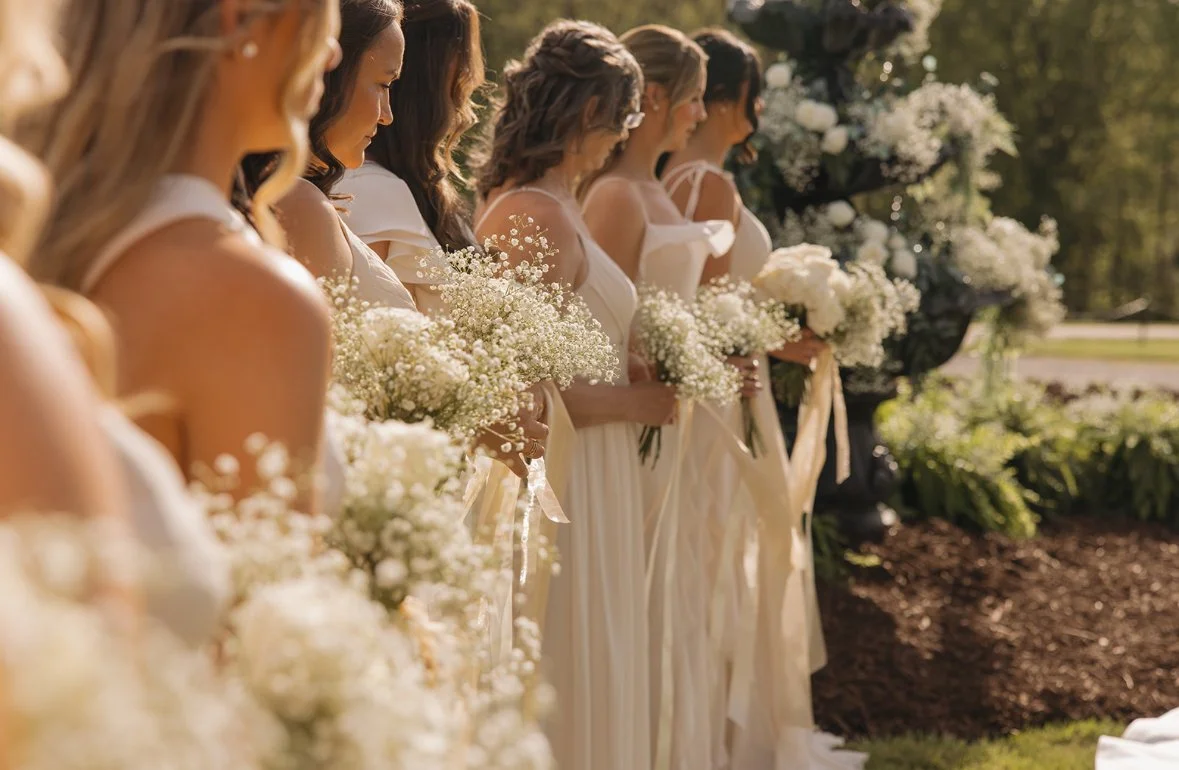 Group of seven women in white dresses holding bouquets of white flowers at an outdoor wedding or event, standing in a line with greenery and flowers in the background.