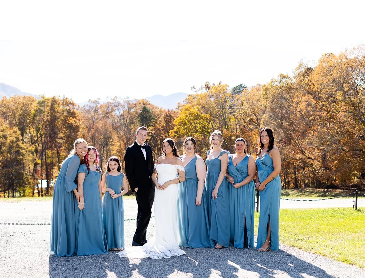 A group of people at a wedding outdoors with fall foliage in the background, including the bride in a white gown, a groom in a black tuxedo, and seven women in matching blue dresses.