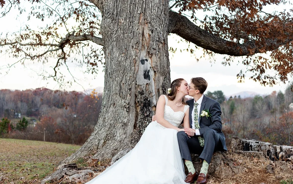 A newlywed couple sharing a kiss under a large, leafless tree in a park during fall.
