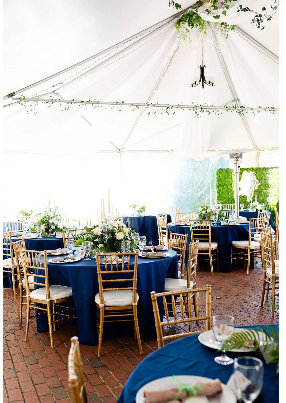 Elegant event setup with round tables draped in navy blue tablecloths, gold chairs with white cushions, and floral centerpieces. The event is indoors with a white canopy overhead decorated with greenery, and brick flooring.