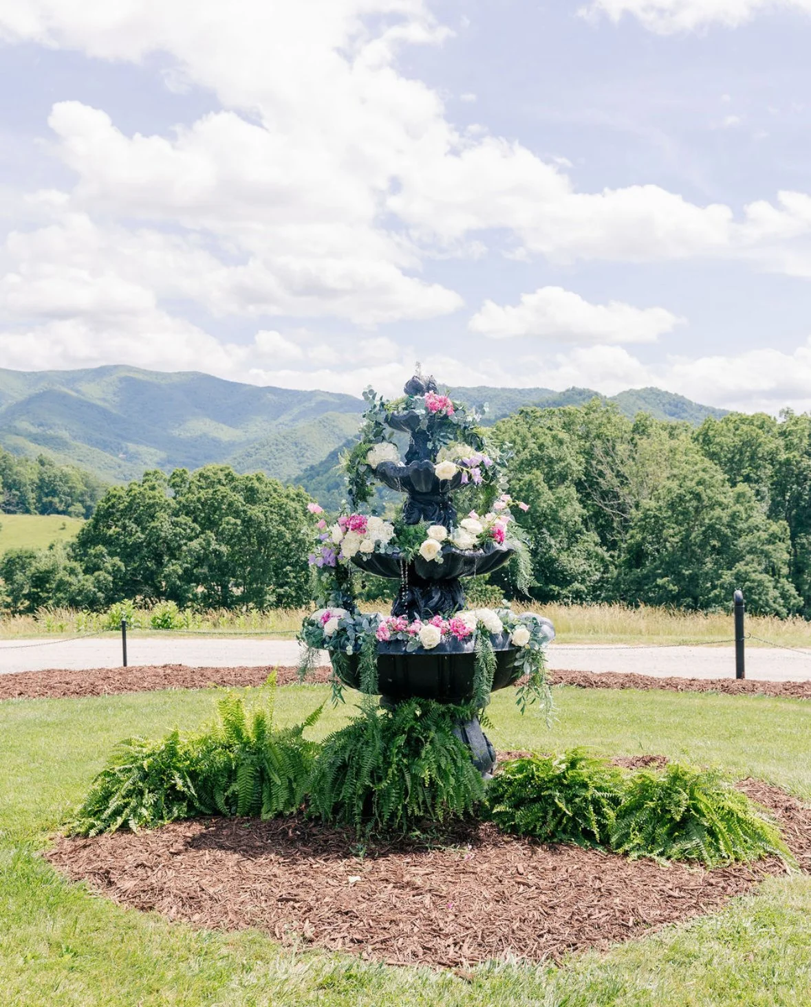 A decorative black fountain adorned with pink, white, and purple flowers, surrounded by green ferns and set in a landscaped garden with mountains and a cloudy sky in the background.