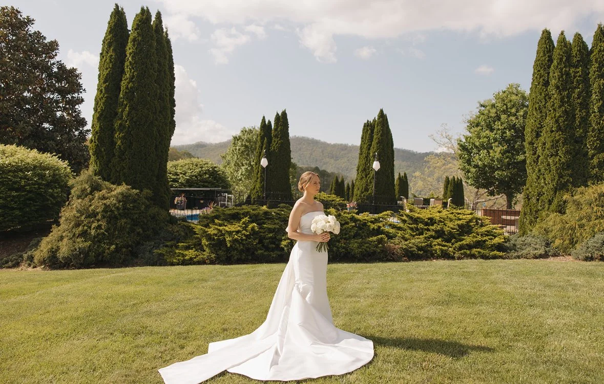 A bride in a white wedding dress stands on a grassy lawn holding a bouquet of white flowers with trees and hills in the background.