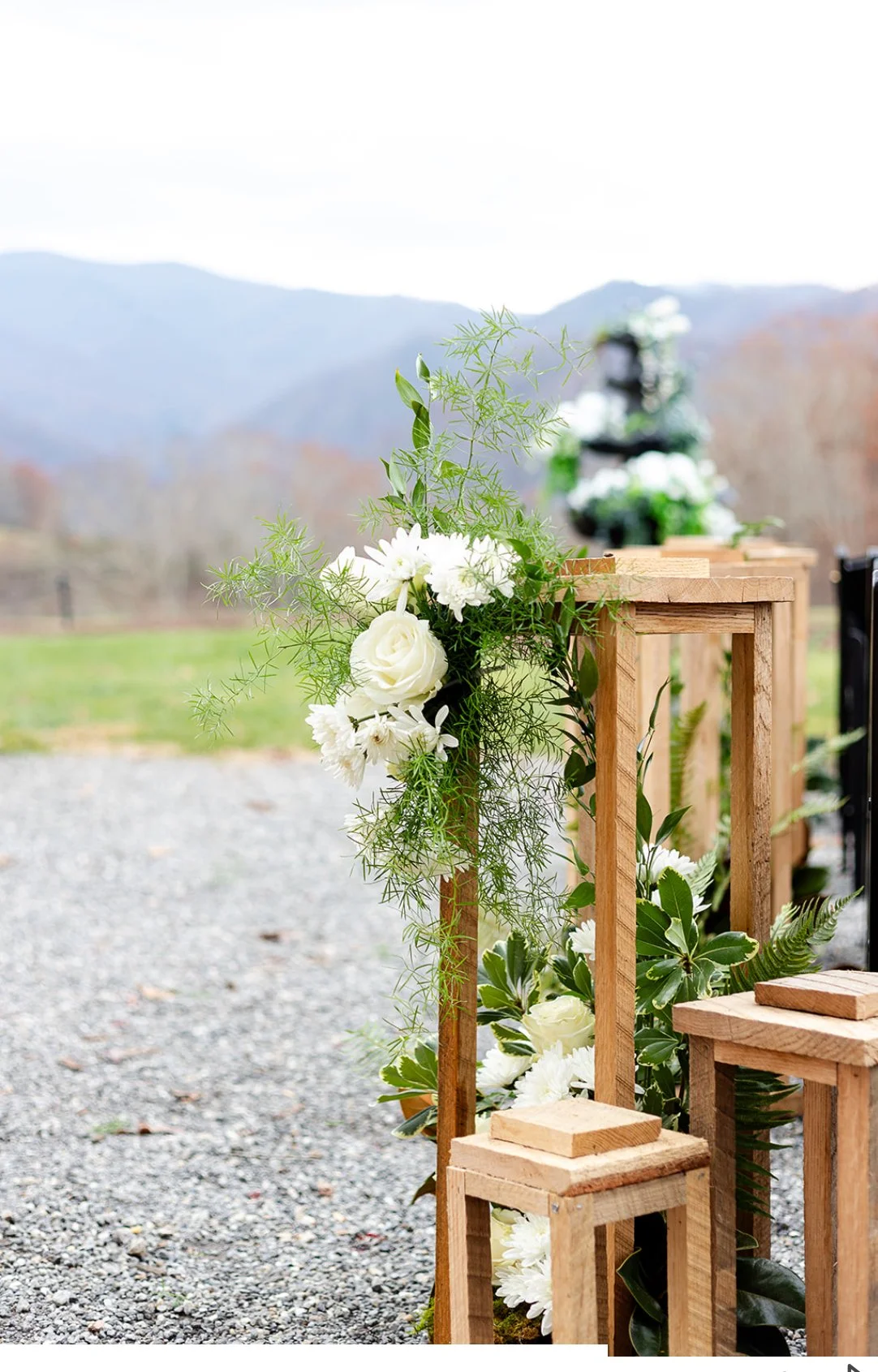 Outdoor wedding setup with wooden chairs decorated with white flowers and greenery, set against a rural mountain background.