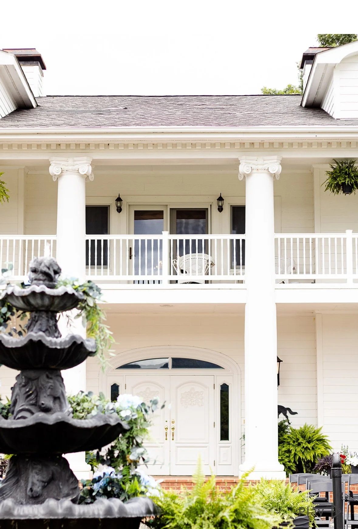 Front entrance of a large white house with tall columns, a second-story balcony with white railing, and hanging plants. There is a decorative black fountain in the foreground surrounded by green plants.