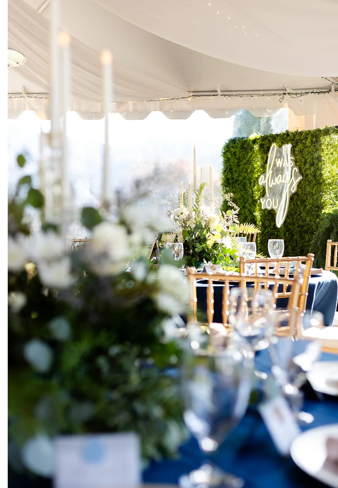 Decorated outdoor wedding reception setup with a table, floral arrangements, glasses, and a green hedge backdrop with illuminated cursive sign that reads "I was always you."