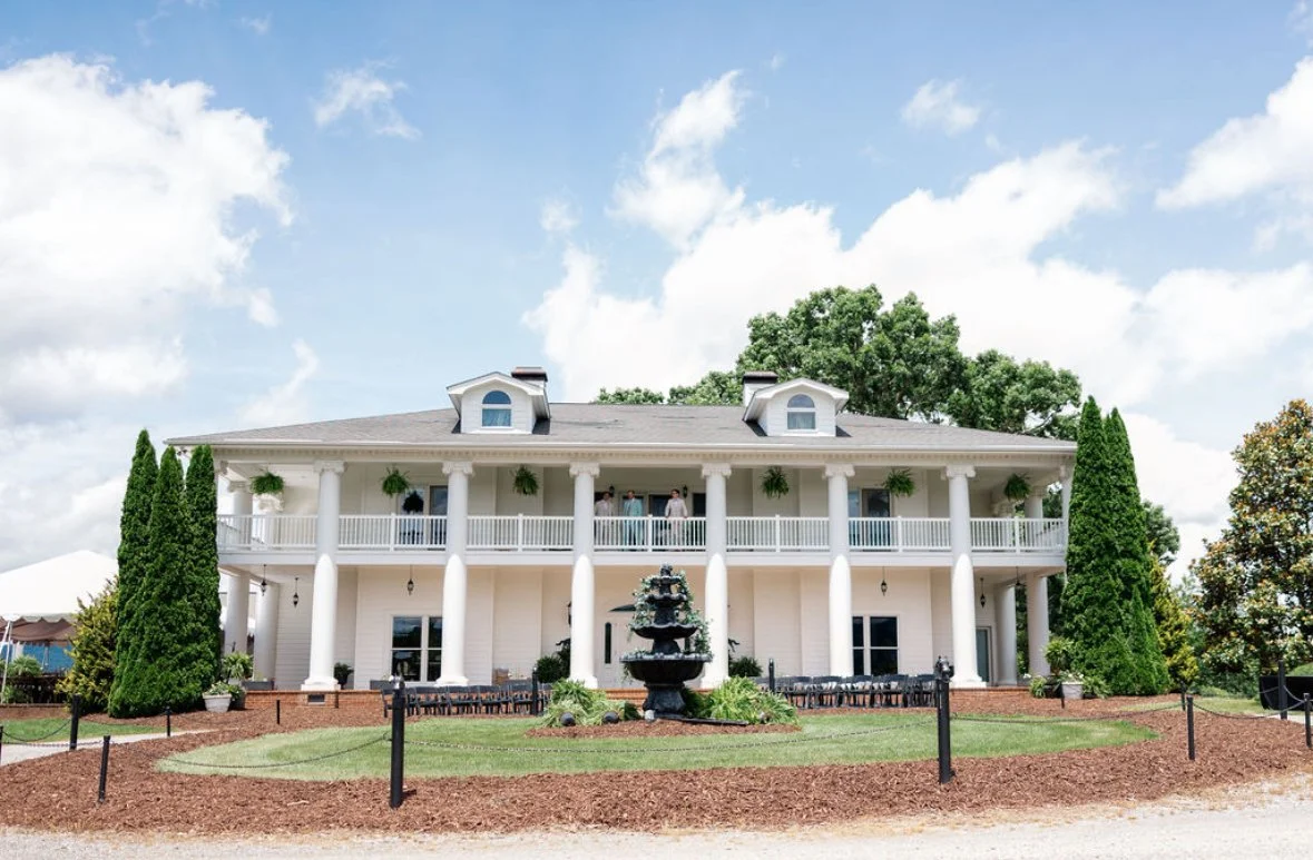Large white two-story house with a spacious balcony, tall trees, and a central fountain in front, under a blue sky with clouds.
