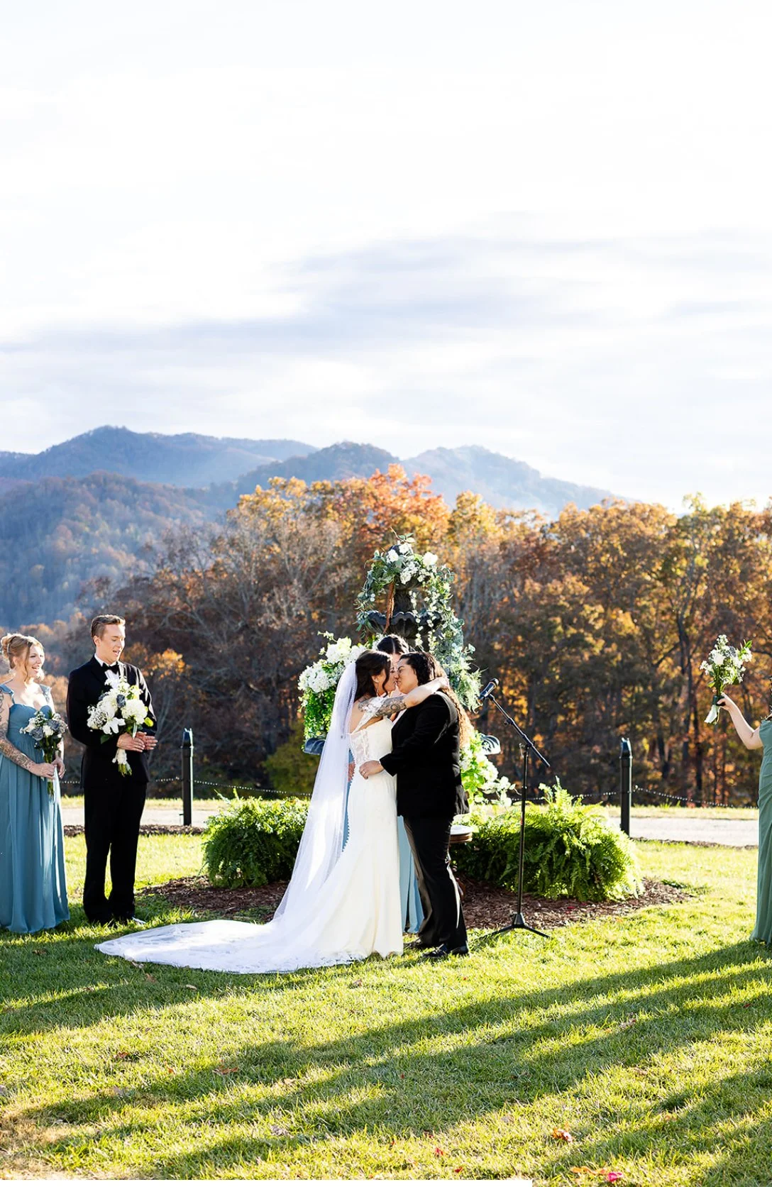 A wedding ceremony outdoors with a bride and groom kissing, surrounded by bridesmaids and groomsmen holding bouquets, with a scenic mountain backdrop and colorful fall trees.