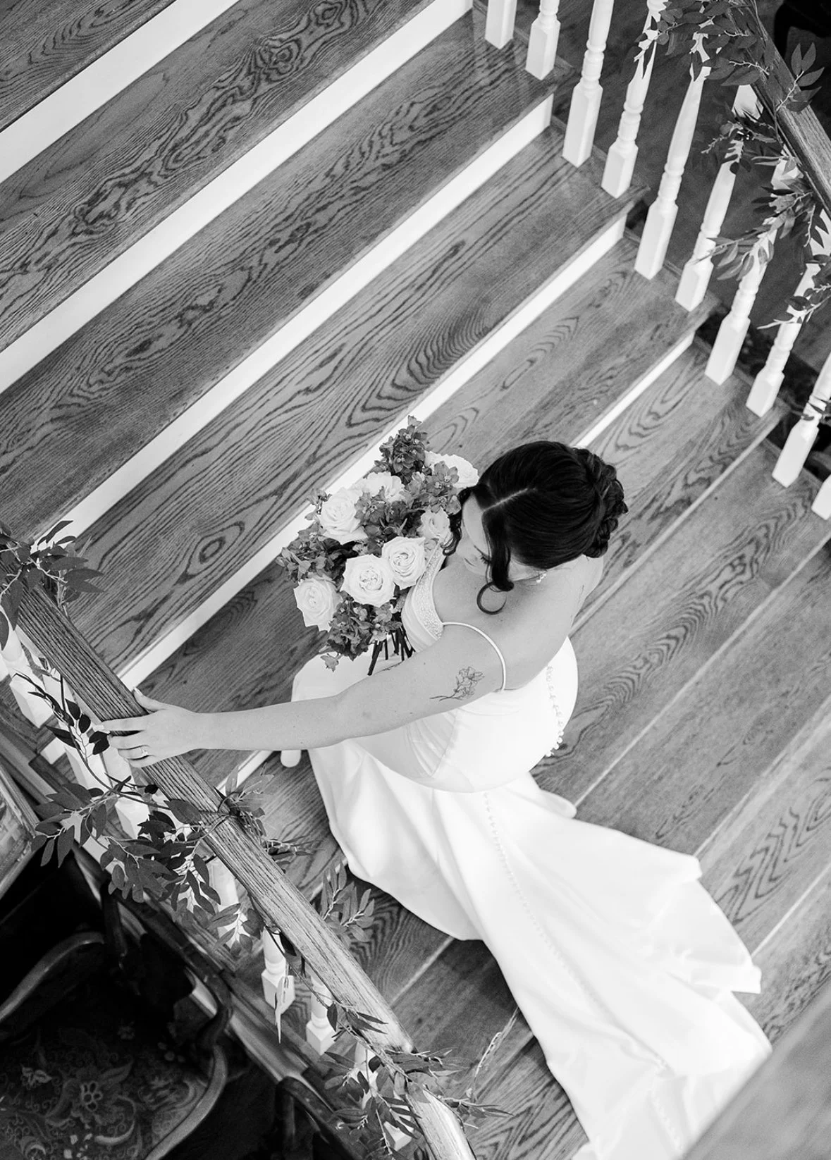 A bride in a wedding dress holding a bouquet of roses and greenery, standing on a wooden staircase decorated with greenery.