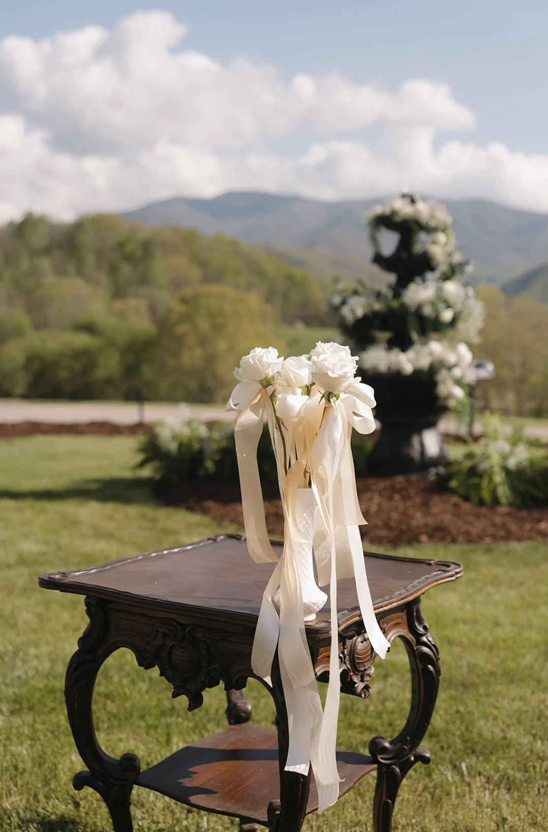 A small vintage wooden table with a bouquet of white roses and ribbons on top, set outdoors with a scenic mountain backdrop and a multi-tiered floral fountain in the background.