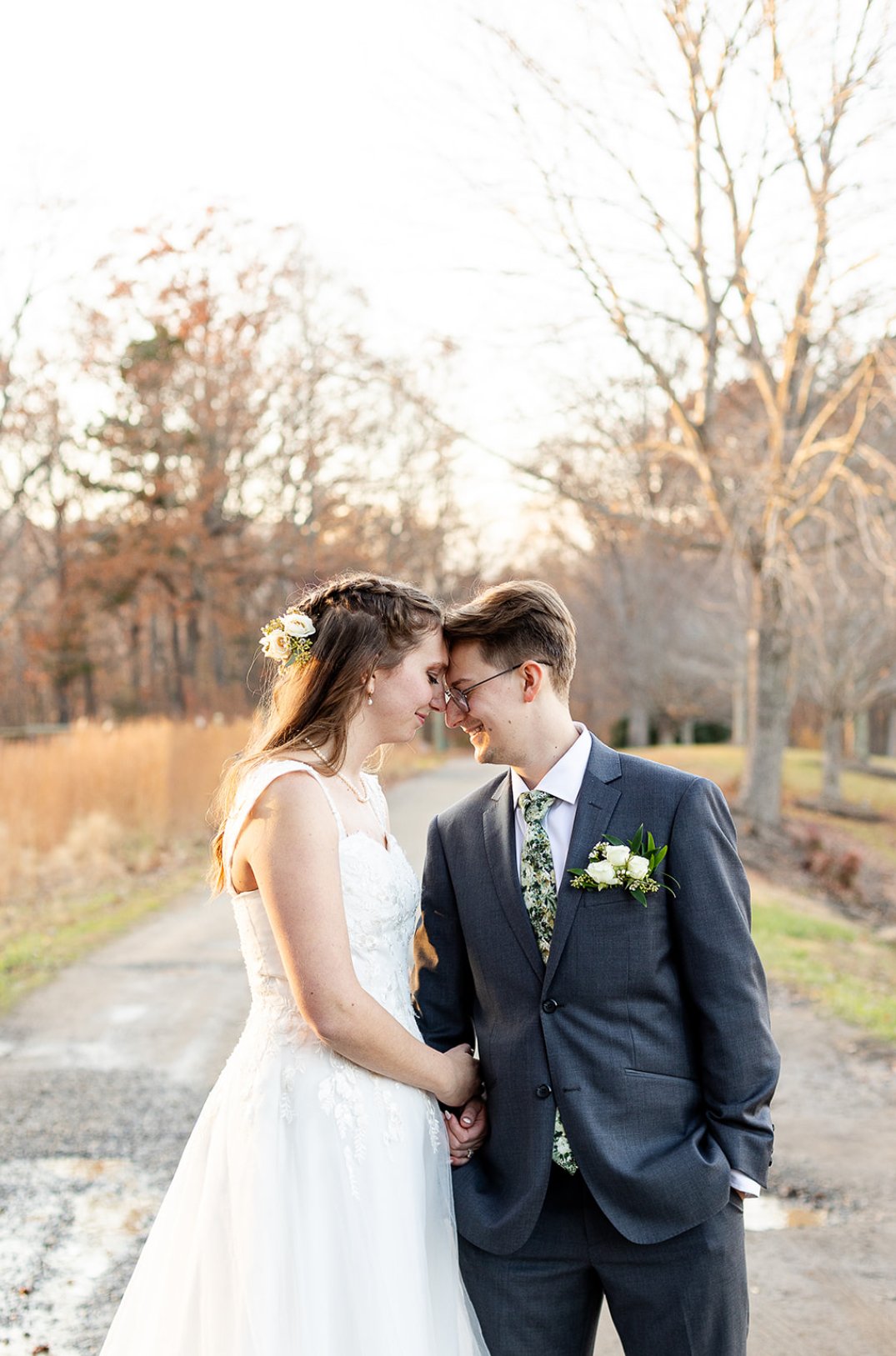 A bride and groom touching foreheads and smiling, holding hands outdoors during sunset, with leafless trees in the background.