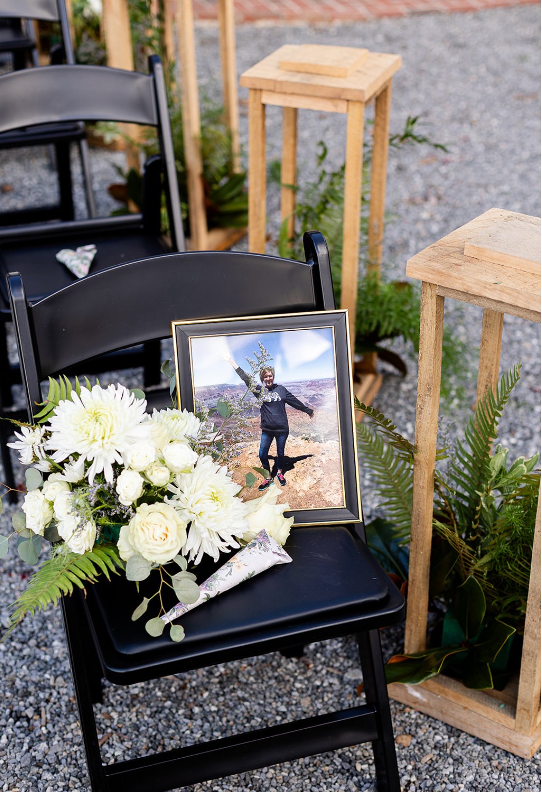 Black chair with a framed photograph of a woman at the Grand Canyon, a bouquet of white flowers, and a small wrapped gift. Two wooden pedestals with plants are in the background.