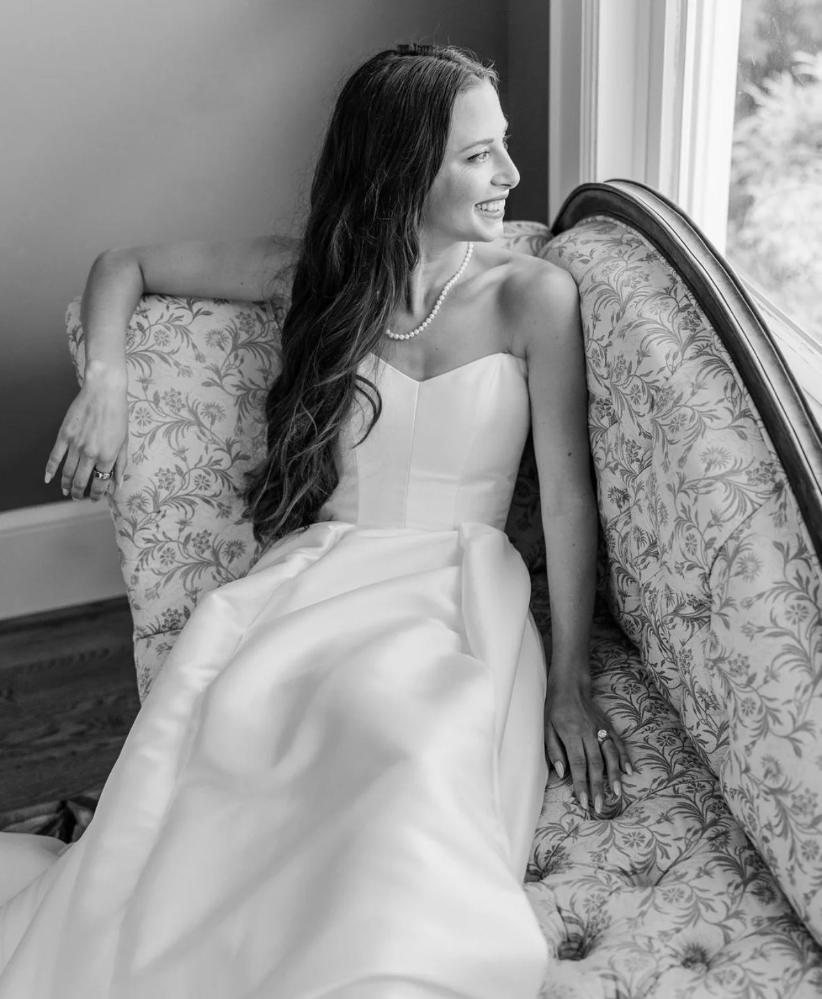 A woman in a wedding dress sitting on a floral upholstered sofa, looking out the window and smiling.