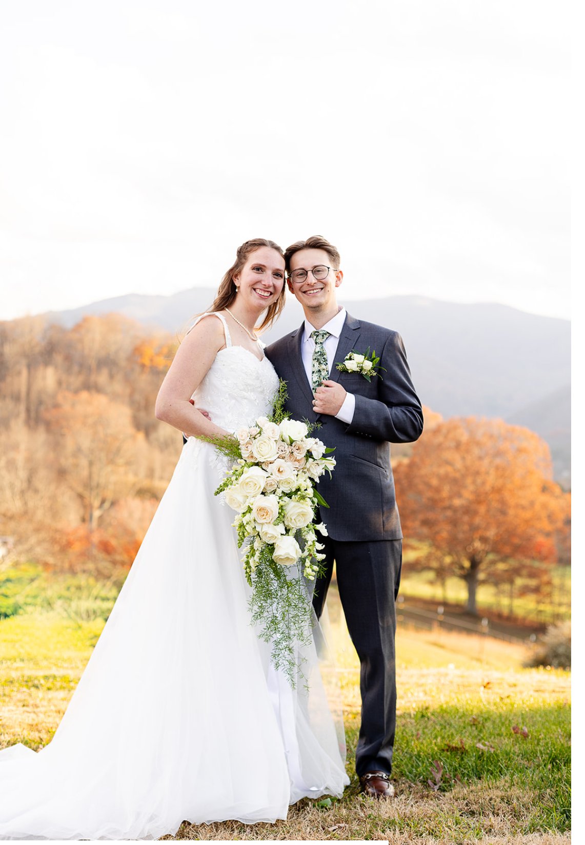 A newlywed couple standing outdoors during fall, smiling, with autumn trees and mountains in the background. The bride is holding a cascading bouquet of white roses and greenery, and both are dressed in wedding attire.