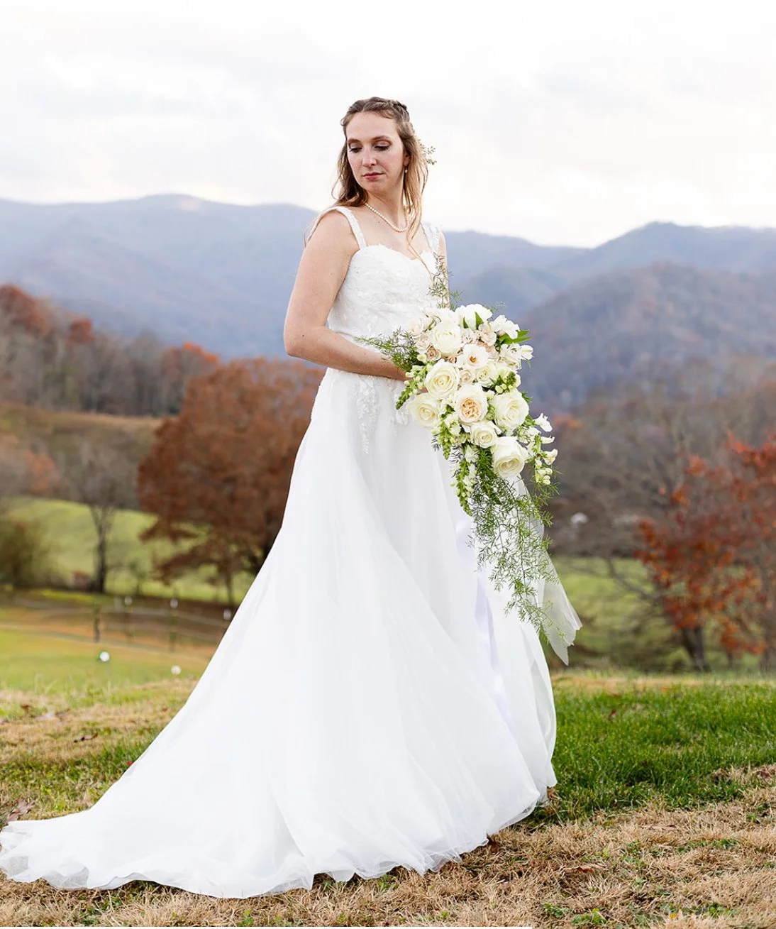 A bride in a white wedding dress holding a cascading bouquet of white roses and greenery, standing outdoors on grass with mountains and autumn trees in the background.