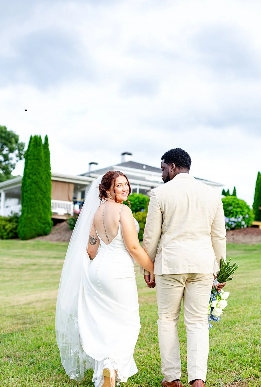 A newlywed couple holding hands and walking on a grassy lawn on a cloudy day, with a house and trees in the background.