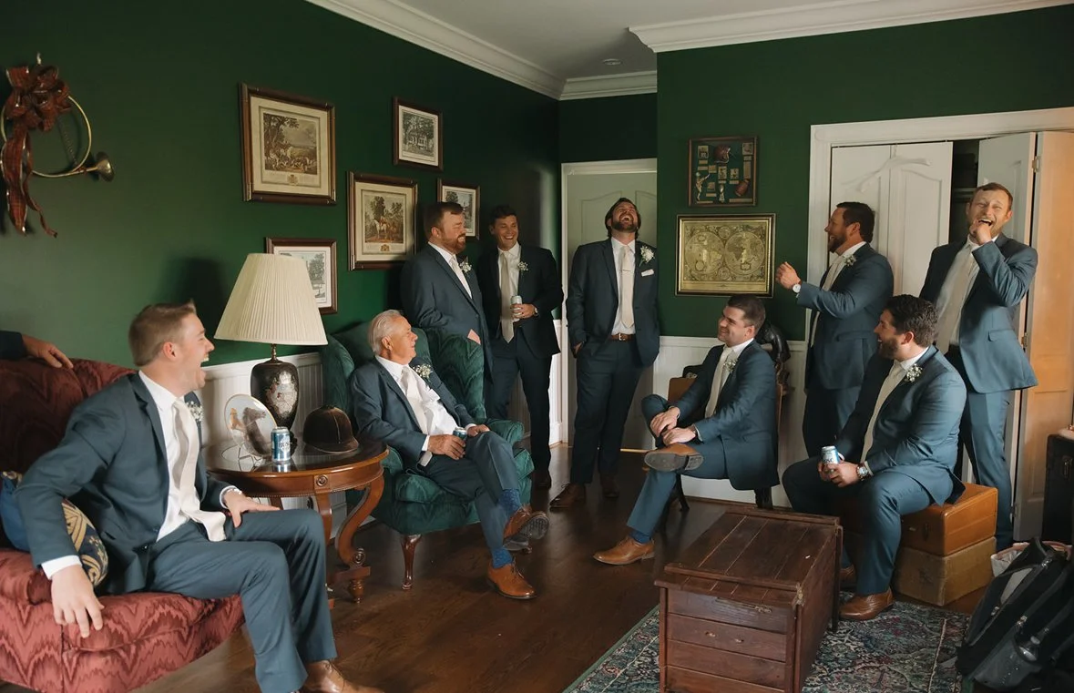 Men in suits gathered in a living room, chatting and laughing, with some sitting and others standing.