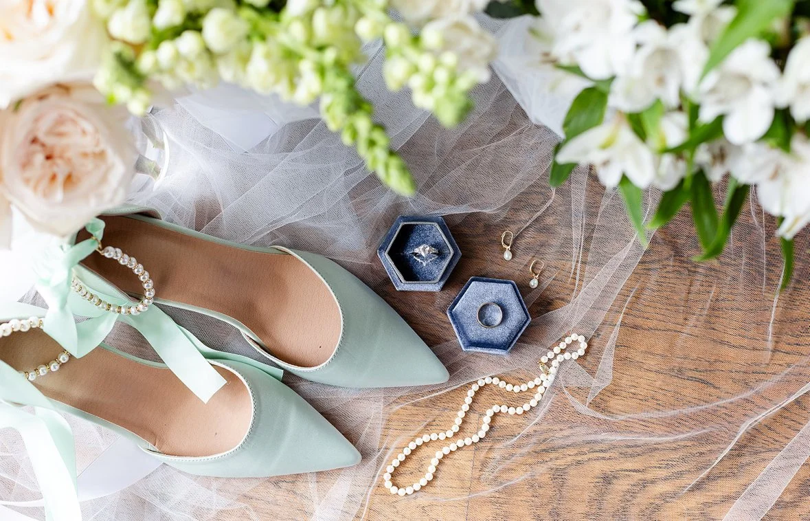 Mint green high heel shoes with ankle straps and pearl embellishments, jewelry including pearl earrings and a pearl necklace, and a diamond ring in a blue velvet box, on a sheer fabric background with flowers.