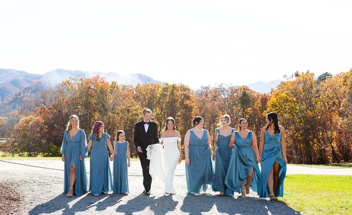 Bride in white dress walking with bridesmaids and a groomsman in black tuxedo outdoors on a sunny day with fall foliage in the background.