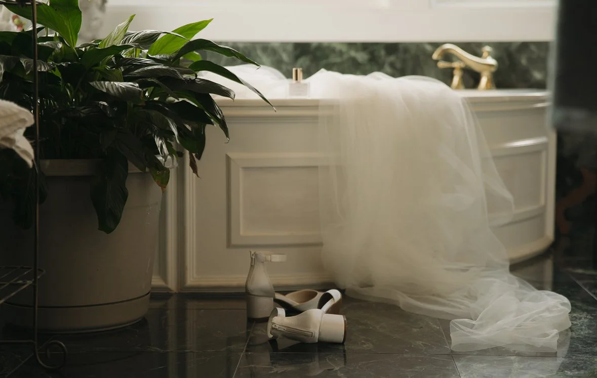 A bathroom with a large potted plant, a white vanity with a gold faucet, and a sheer fabric draped over the countertop. There are a bottle of soap, a shoe, and a shoehorn on the dark marble floor.
