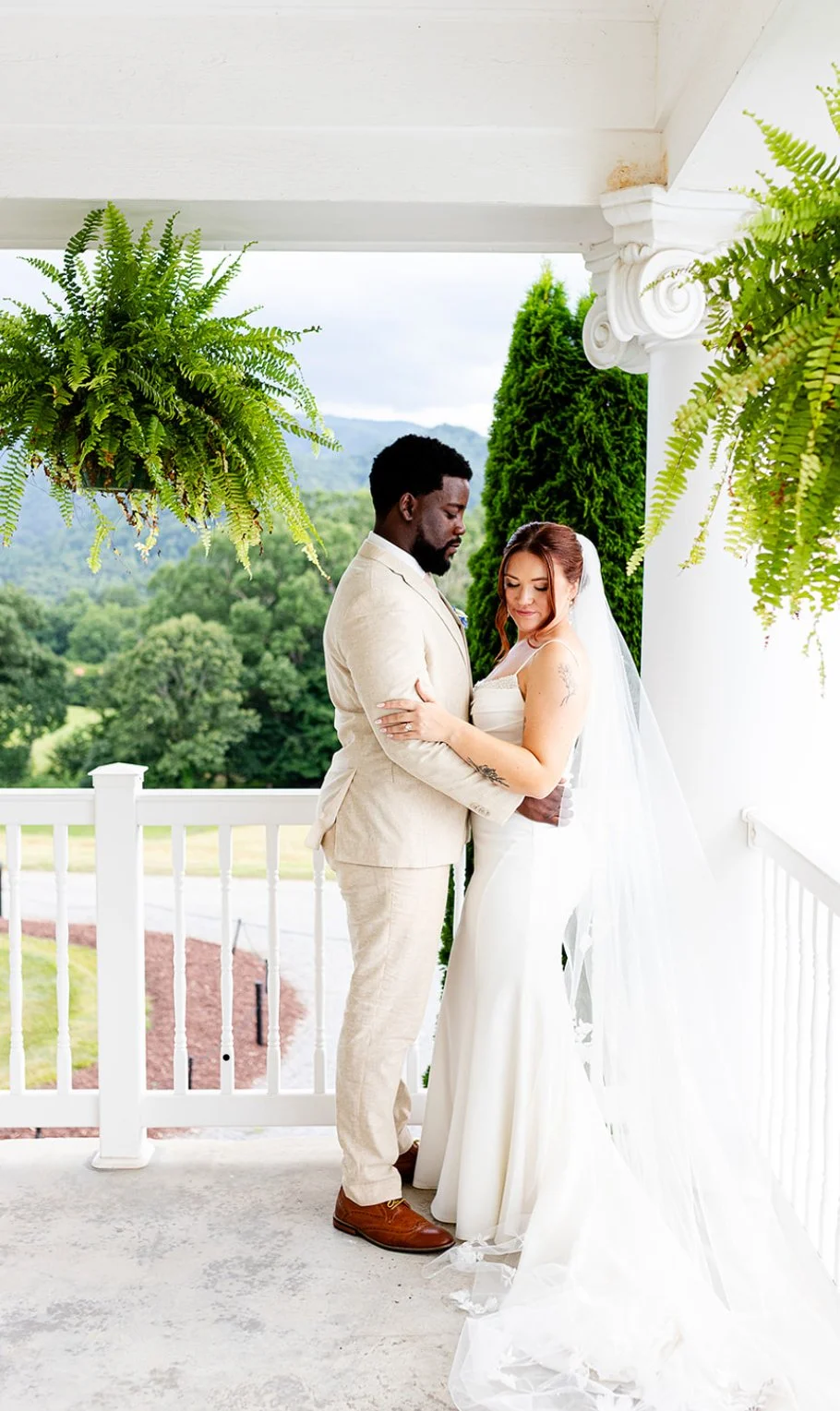 A bride and groom standing close together on a white porch with green trees and mountains in the background, dressed in wedding attire.