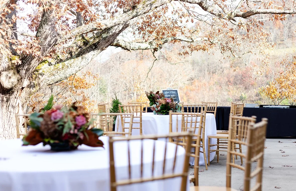 Outdoor event setup with round tables draped in white tablecloths, decorated with floral centerpieces, and surrounded by wooden chairs, under a large tree with autumn-colored leaves.
