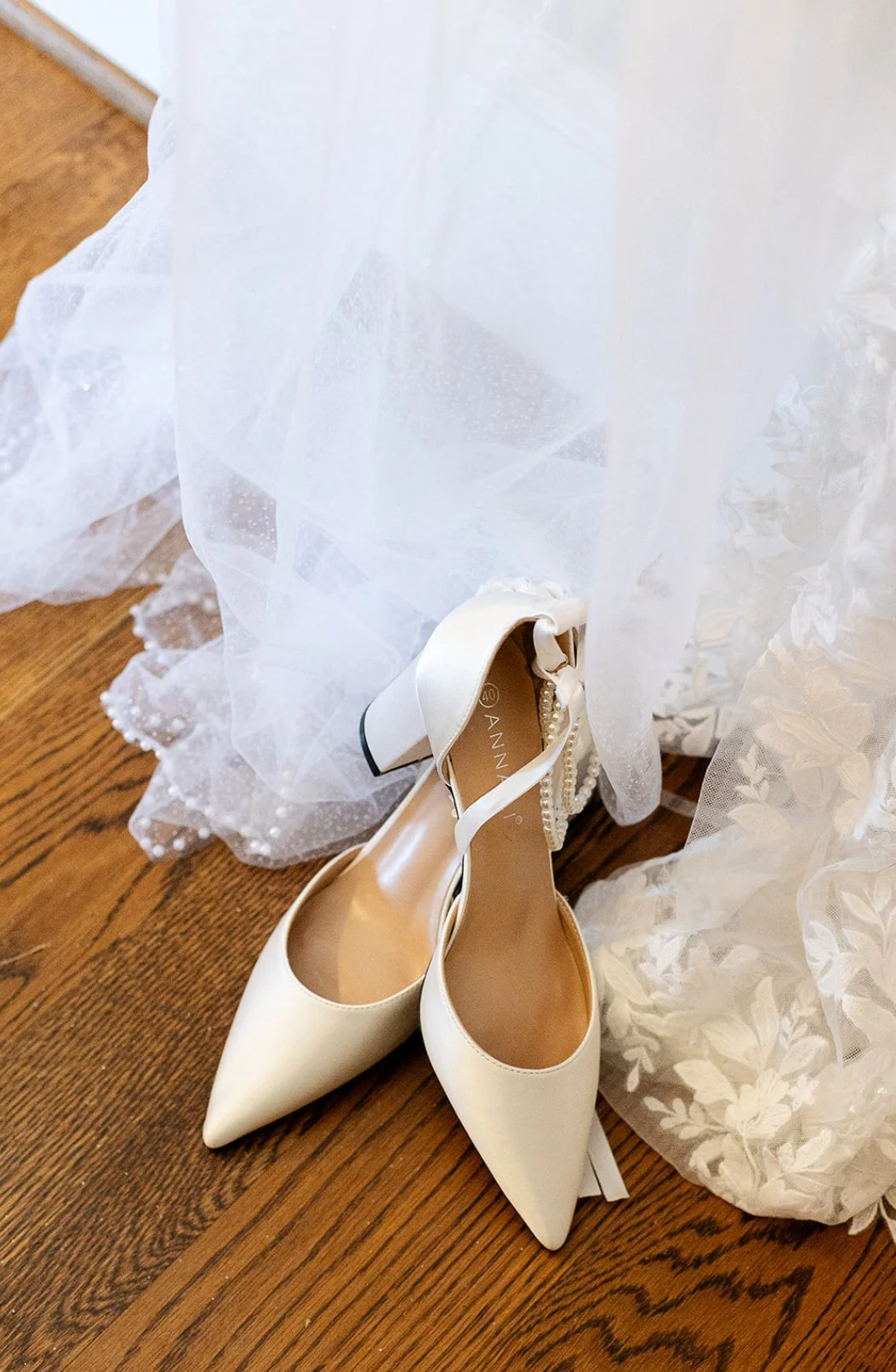 A pair of white high-heeled shoes with pointed toes resting on a wooden floor, partially covered by a white, lace wedding dress.
