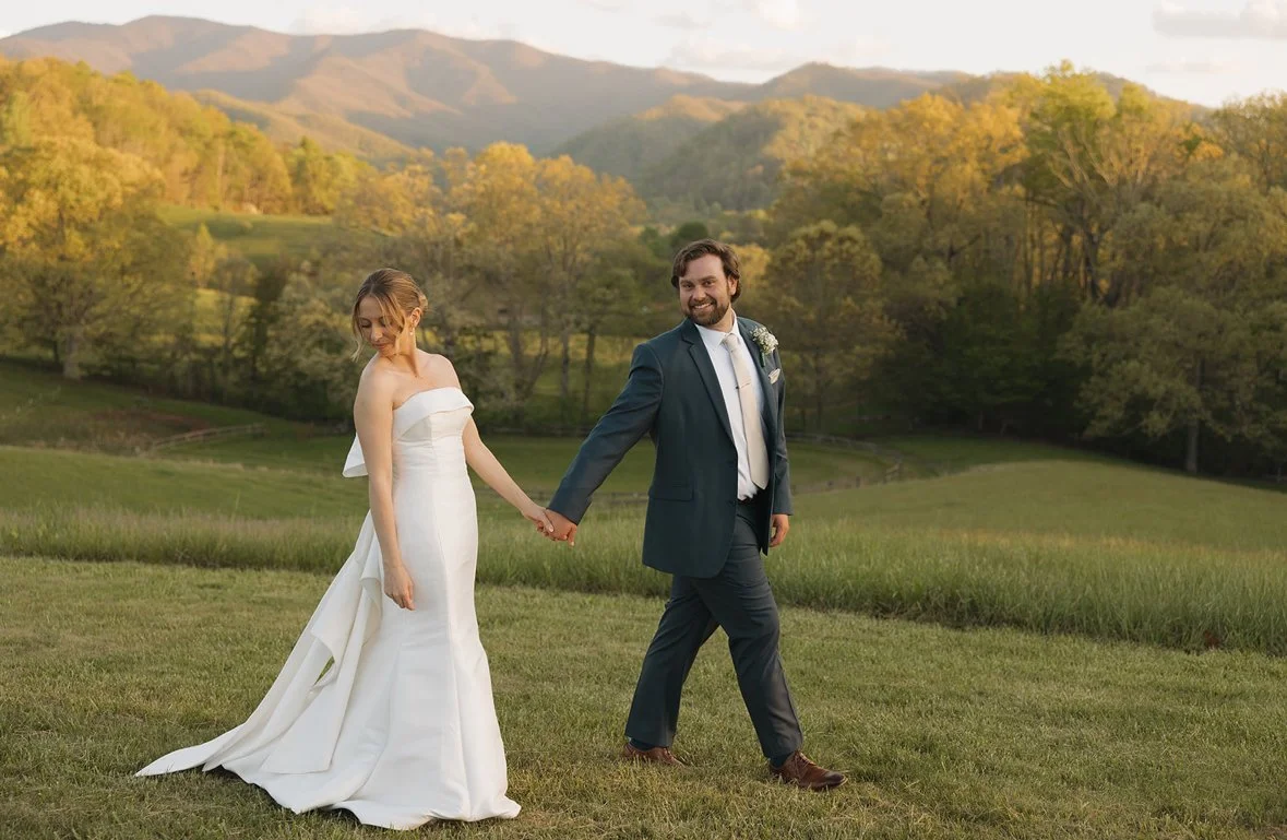 A bride and groom holding hands in a field with mountains and trees in the background during sunset.