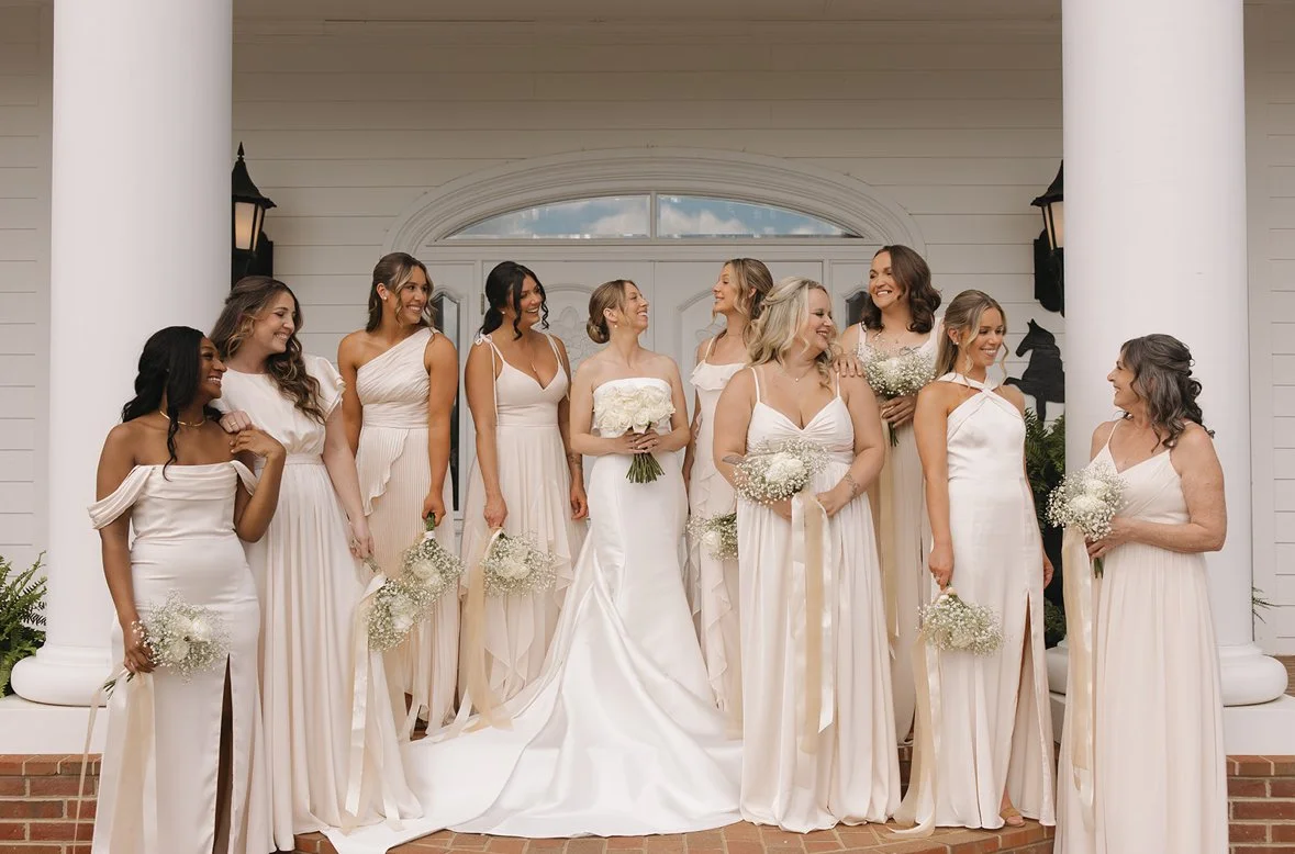 A group of women dressed in white wedding gowns standing on the porch of a house, smiling and looking at each other, holding bouquets.