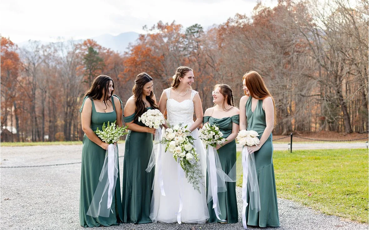 A bride and four bridesmaids standing outdoors on a gravel path during fall, holding bouquets and smiling at each other with trees in the background.