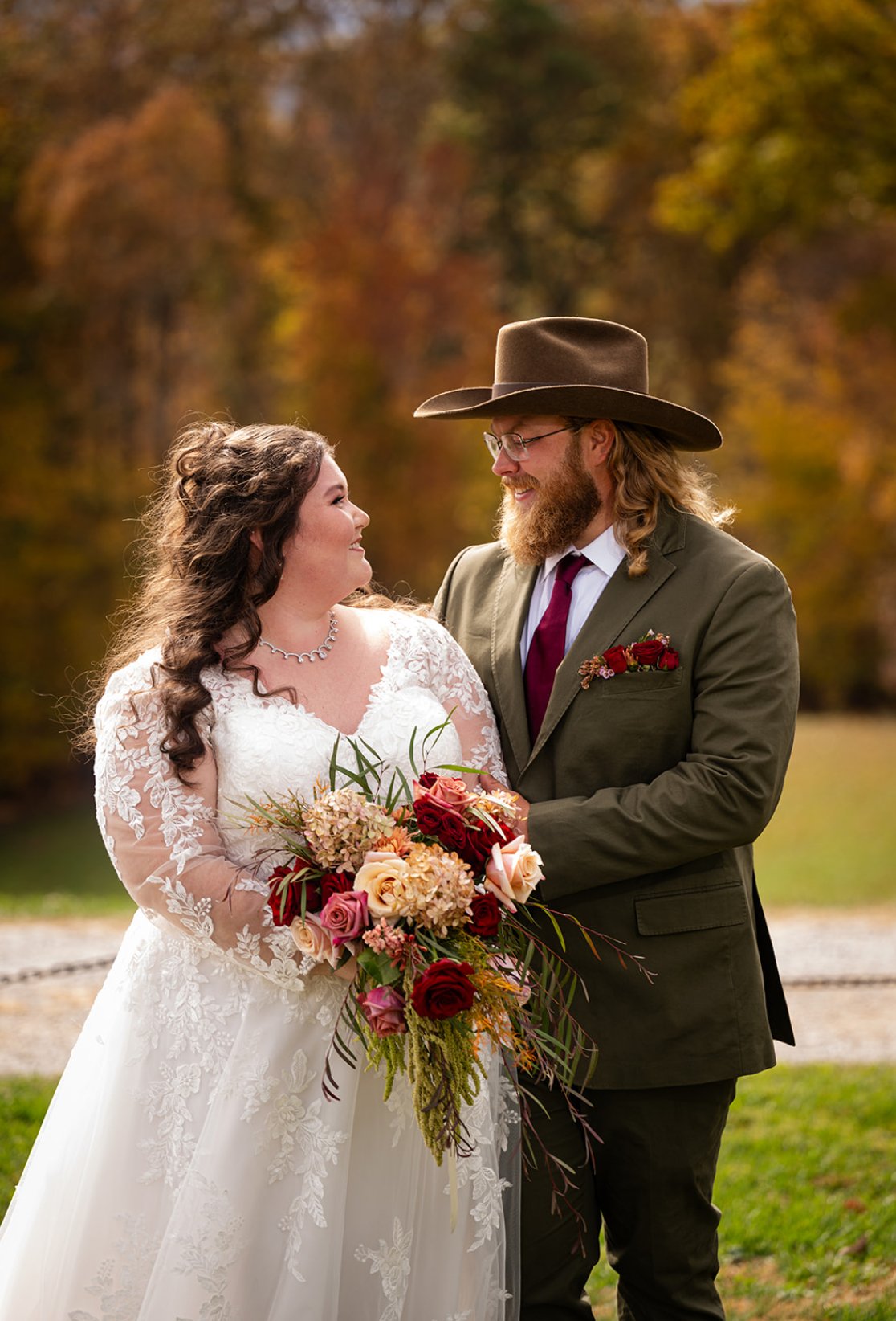A bride and groom facing each other outdoors during fall, with colorful trees in the background. The bride is wearing a white lace wedding dress and holding a large bouquet of pink, red, and cream flowers. The groom is dressed in a green suit with a 