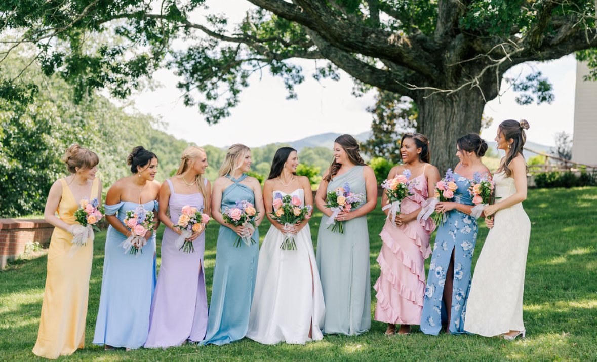 Group of women in colorful dresses holding bouquets of flowers standing outdoors under a large tree.