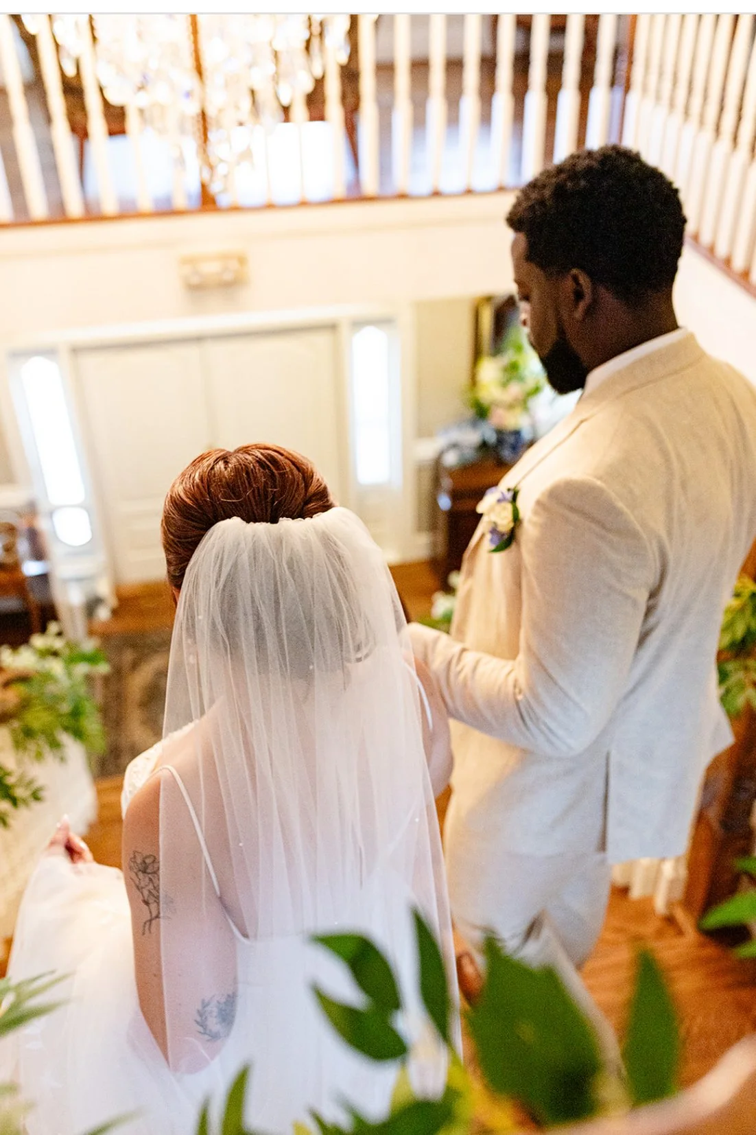 A bride and groom exchanging vows during a wedding ceremony indoors, with the bride wearing a veil and a white dress, and the groom in a cream-colored suit.