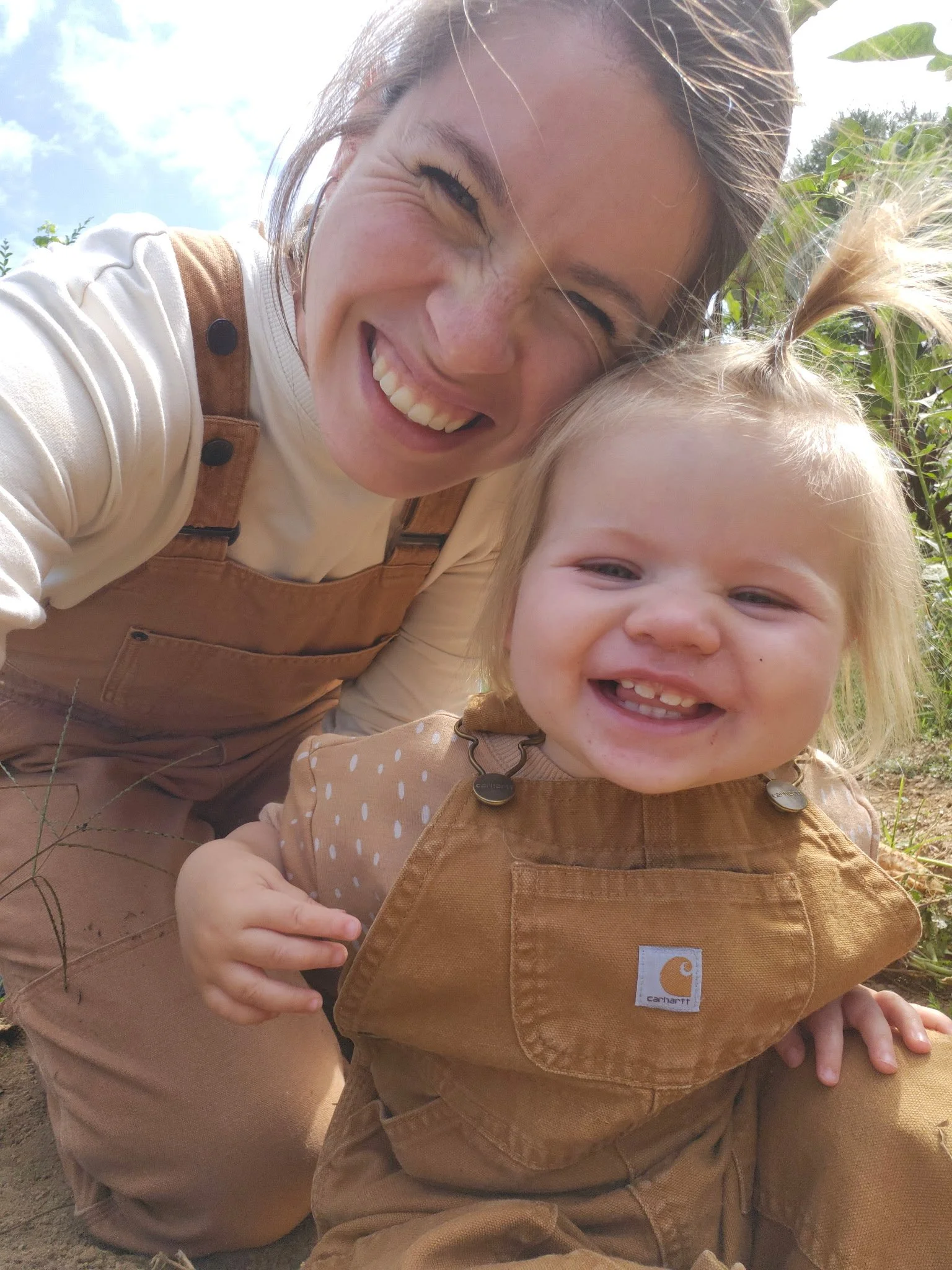 A woman and a young girl smiling, close together outdoors on a sunny day.