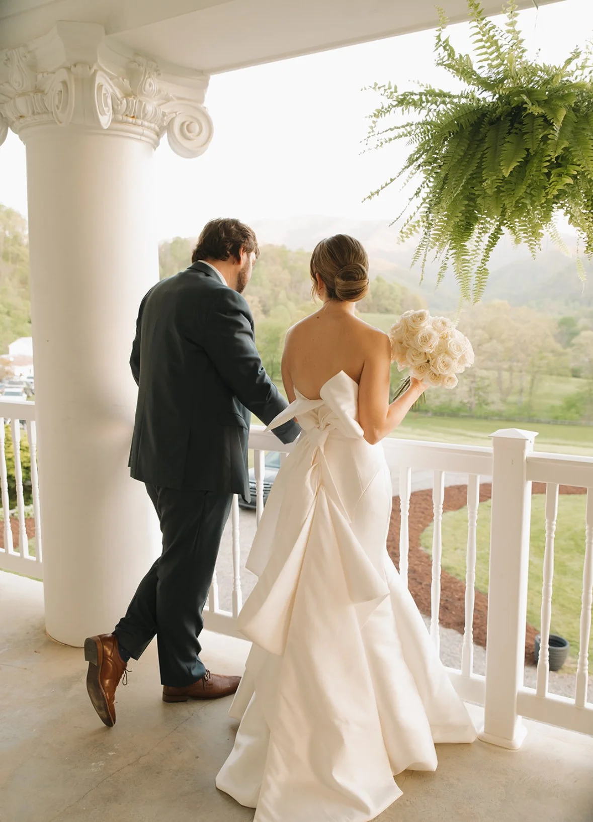 A bride and groom standing on a porch, facing away, with the bride holding a bouquet of white roses. The groom is dressed in a black suit, and the bride is wearing a strapless white wedding gown with a large bow. A lush, green landscape is visible ou