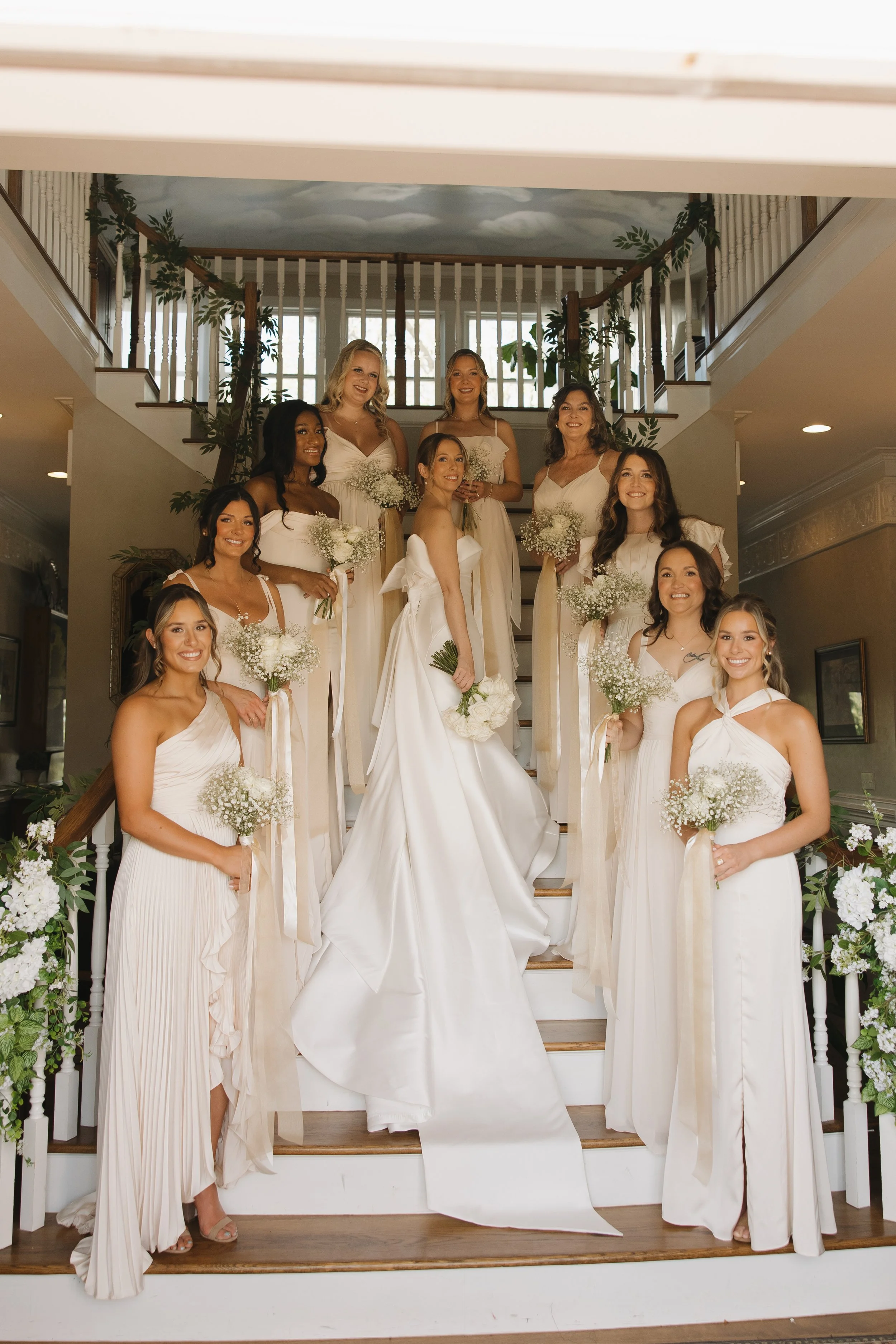 A bride with her bridesmaids on a staircase decorated with white flowers and greenery during a wedding celebration.