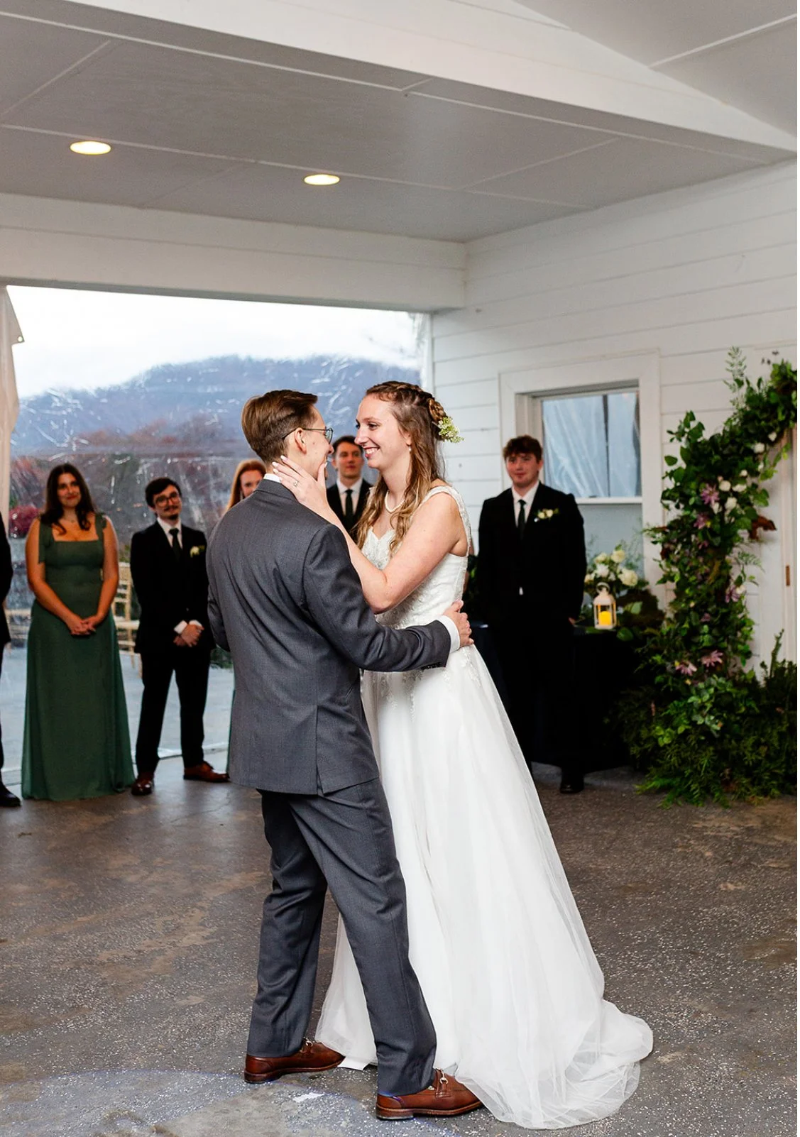 A bride and groom sharing their first dance inside a wedding venue with mountain views outside. The bride is smiling and holding the groom's face, while the groom looks at her. Bridesmaids and groomsmen are watching in the background.