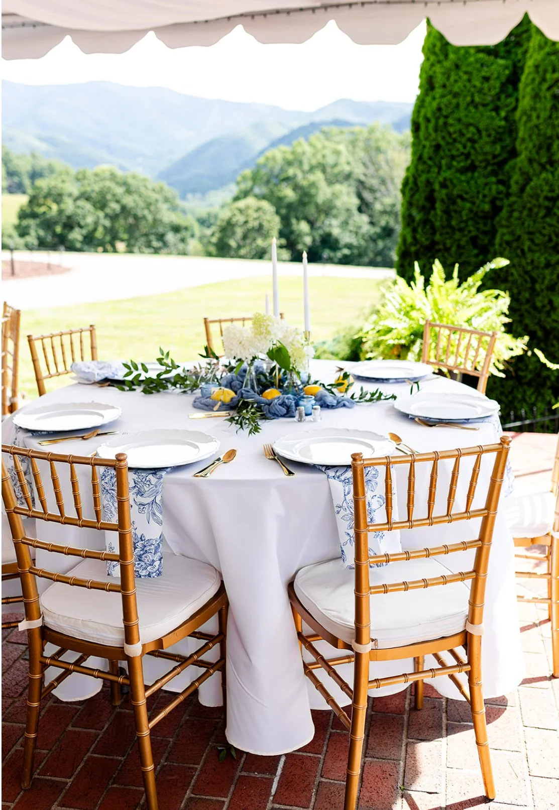 Outdoor dining table set with white tablecloth, gold flatware, and white plates, decorated with a floral centerpiece and lemons, on a brick patio with a scenic mountain view.
