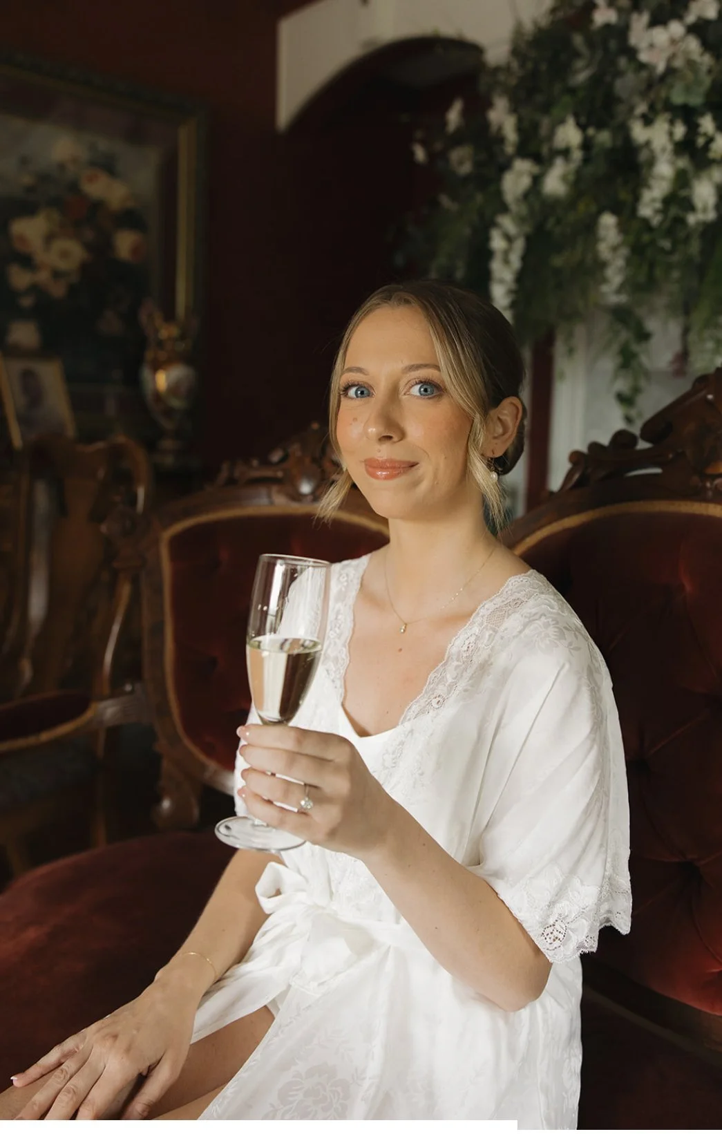 A woman in a white lace dress sitting on a vintage red velvet couch, holding a glass of champagne, in a richly decorated room with floral arrangements and classic artwork.