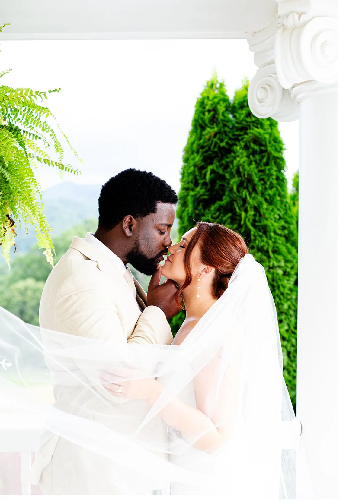 A bride and groom sharing a kiss outdoors, with greenery and trees in the background, white architectural columns, and a flowing veil.