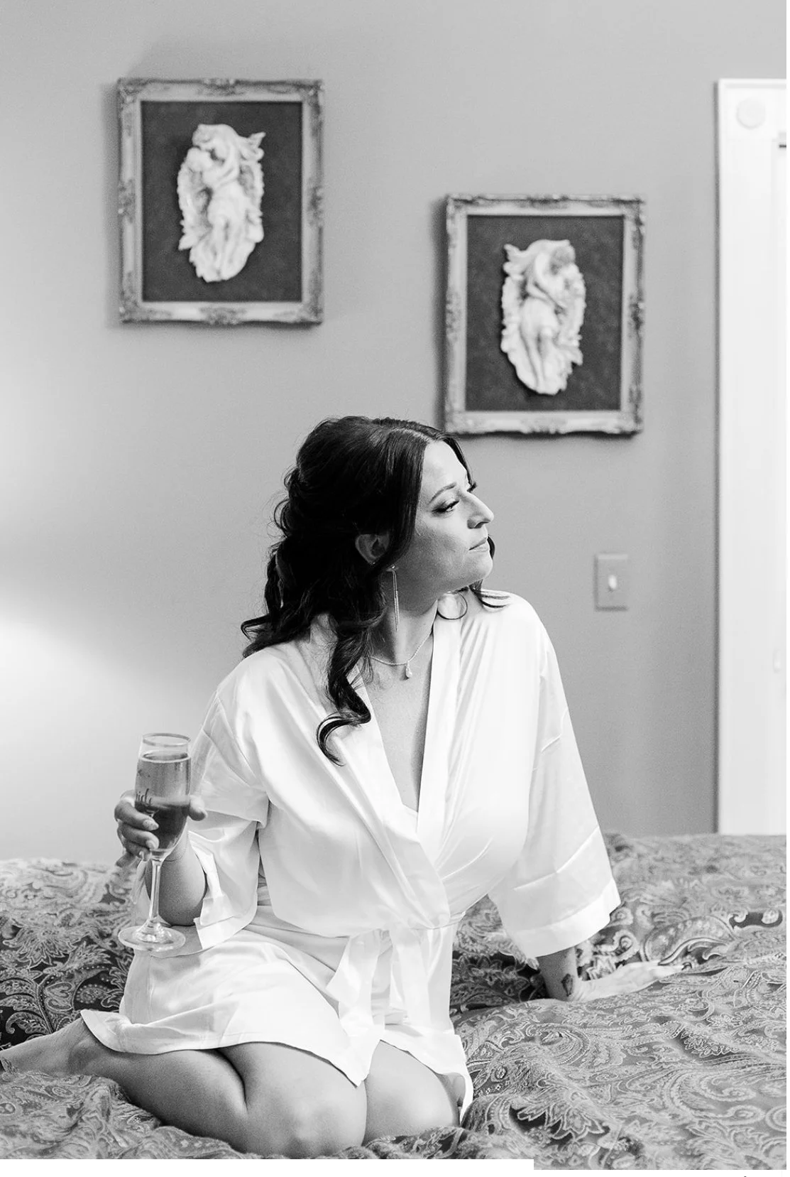A woman in a white robe sitting on a bed holding a glass of champagne. She has dark wavy hair and looks to the side with a serene expression. Behind her are two framed relief sculptures of horses on the wall. The photo is in black and white.